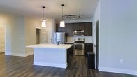 This single-floor space is designed with a clean, contemporary aesthetic that emphasizes both function and style. The open-concept kitchen serves as the heart of the unit, anchored by a spacious white-topped island with an integrated sink and pendant lighting above, making it ideal for both meal preparation and casual dining. Dark espresso-toned cabinetry lines the kitchen walls, offering ample storage while providing a striking contrast against the light-colored walls and countertops. The kitchen is fully equipped with a stainless steel refrigerator, electric range, and built-in microwave, giving it a polished, move-in-ready appeal. A track lighting system overhead ensures the cooking area is well-lit and practical. Rich, dark hardwood-style flooring runs throughout the space, adding warmth and continuity to the open layout. The overall color palette — combining dark cabinetry, white surfaces, and neutral walls — creates a balanced and inviting atmosphere. Additional doors visible from the main area suggest connectivity to separate rooms, contributing to a functional and well-organized floor plan. The space is thoughtfully designed to maximize usability while maintaining a modern, cohesive visual identity.