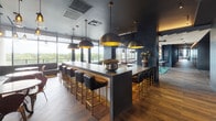 Located in Houston, TX 77024, this upscale single-floor amenity lounge is a masterclass in contemporary interior design, blending dark, moody aesthetics with warm wood accents and gold-toned hardware throughout. The space is organized into several distinct zones, each thoughtfully curated for social and recreational use.

The bar area features a sleek concrete-topped island surrounded by quilted leather barstools with brass legs, illuminated by large brass dome pendant lights. A mirrored back wall and mounted flat-screen televisions complete the entertainment-ready atmosphere, while wood-look tile flooring in a herringbone pattern adds texture underfoot.

A bright lounge area opens to sweeping floor-to-ceiling windows and glass doors that frame lush treetop views, flooding the space with natural light. Leather sofas and accent chairs are arranged around low coffee tables beneath sculptural pendant lighting, creating an inviting setting for relaxed gatherings.

A dramatic private lounge and screening room features an all-dark interior with a striking slatted wood-and-black ceiling, built-in banquette seating, a long communal bar table with black-and-brass stools, and a large wall-mounted display screen — ideal for private events or watch parties.

A long corridor connects the zones, accented by exposed beam ceilings, round copper-framed mirrors, and bar cart vignettes that reinforce the space's refined, hospitality-inspired character.