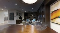 Located at 201 Willett Ave in Port Chester, NY, this single-floor commercial space presents a polished and contemporary interior designed to impress. The main lounge area serves as the focal point, featuring a carefully curated arrangement of plush, modern armchairs accented with teal throw pillows, centered around a dark wood coffee table atop a patterned area rug. The space is anchored by a reception or service counter along one wall, offering a functional yet stylish point of contact. Ambient and recessed lighting work in harmony with a striking globe pendant light to create a warm, inviting atmosphere despite the predominantly dark color palette of charcoal and deep brown tones. A feature wall with geometric cutouts and built-in shelving adds architectural depth and visual interest. Decorative elements, including framed wall art, sculptural wire accent pieces, and an ornate golden mirror, lend an artistic character to the space. Workstation areas with monitors are subtly integrated into the background, suggesting a multifunctional use. Rich hardwood flooring grounds the design, while the overall layout promotes both comfort and professionalism, making it well-suited for a hospitality, corporate, or mixed-use environment.