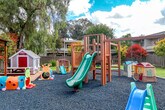 Located at 1901 Halford Ave in Santa Clara, California, this thoughtfully designed outdoor space offers a well-rounded environment for play, relaxation, and recreation. The centerpiece of the grounds is a generously sized children's playground area, surfaced with blue rubber mulch for safety. It features a multi-level wooden climbing structure with slides, a charming playhouse, and an assortment of smaller play equipment, all arranged to encourage active outdoor engagement. Adjacent to the playground, a redwood-toned pergola provides a shaded seating area with benches, offering a comfortable retreat for supervision or relaxation. A wide open lawn of lush green turf extends across the property, bordered by mature trees including tall weeping varieties that provide natural shade and privacy. The landscaping is well-maintained throughout, with decorative rock arrangements, colorful Japanese maples, ornamental shrubs, and seasonal plantings lining the perimeter fencing. A covered gazebo structure on one side of the property houses additional equipment, adding further utility to the space. The fully fenced grounds ensure a secure and contained environment. Abundant natural light and the surrounding tree canopy create a pleasant, park-like ambiance throughout the entire outdoor area.