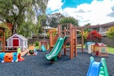 Located at 1901 Halford Ave in Santa Clara, California, this thoughtfully designed outdoor space offers a well-rounded environment for play, relaxation, and recreation. The centerpiece of the grounds is a generously sized children's playground area, surfaced with blue rubber mulch for safety. It features a multi-level wooden climbing structure with slides, a charming playhouse, and an assortment of smaller play equipment, all arranged to encourage active outdoor engagement. Adjacent to the playground, a redwood-toned pergola provides a shaded seating area with benches, offering a comfortable retreat for supervision or relaxation. A wide open lawn of lush green turf extends across the property, bordered by mature trees including tall weeping varieties that provide natural shade and privacy. The landscaping is well-maintained throughout, with decorative rock arrangements, colorful Japanese maples, ornamental shrubs, and seasonal plantings lining the perimeter fencing. A covered gazebo structure on one side of the property houses additional equipment, adding further utility to the space. The fully fenced grounds ensure a secure and contained environment. Abundant natural light and the surrounding tree canopy create a pleasant, park-like ambiance throughout the entire outdoor area.