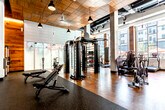 This expansive fitness center is thoughtfully designed to support a full range of workout styles and training needs. The main gym floor features high ceilings with a warm wood slat finish, large pendant lighting, and oversized ceiling fans that create an open, industrial-chic atmosphere. Structural columns and exposed ductwork add to the loft-like aesthetic, while floor-to-ceiling windows along one wall flood the space with abundant natural light and offer views of the surrounding urban landscape.

The strength training area is outfitted with a multi-functional cable machine, weight benches, and dedicated rubber flooring for functional fitness. A mirrored free weight section houses an extensive rack of dumbbells and is accented by white brick walls and a mounted display screen. The cardio zone lines the window wall, featuring a row of treadmills, ellipticals, stair climbers, and rowing machines, all positioned to take advantage of the outdoor views and natural light.

A separate studio space with light wood-look flooring, soaring double-height ceilings, and floor-to-ceiling windows offers a bright, open environment ideal for yoga, stretching, or group classes. Adjacent to it is a vibrant spin and group fitness studio, distinguished by a bold, colorful mural accent wall, ballet barre, mirrored walls, and a large display screen. Private rooms accessible via dark-paneled doors round out the facility.