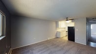 This single-floor space offers a straightforward, functional layout that combines the living and kitchen areas in an open-concept arrangement. The living area is finished with wall-to-wall carpet, providing a comfortable and quiet underfoot feel, while neutral gray walls create a clean, versatile backdrop suited to a variety of furnishing styles. A textured popcorn ceiling runs throughout, adding a classic residential character to the space.

The kitchen is seamlessly integrated at the far end of the living area, featuring white cabinetry that provides ample storage, a sink, and a dishwasher. A black refrigerator anchors one side of the kitchen, offering a subtle contrast to the lighter cabinetry. The kitchen floor transitions to hard surface flooring, clearly delineating the two zones within the open layout.

A ceiling fan with a built-in light fixture is positioned in the kitchen area, providing both air circulation and overhead lighting. A baseboard heater runs along the lower wall, ensuring efficient climate control throughout the space. The overall ambiance is clean, bright, and practical, making this a well-organized space that balances everyday living and functional utility in a compact, single-level footprint.