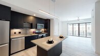 Located at 5211 Peachtree Blvd in Chamblee, Georgia, this single-floor unit showcases a clean, contemporary design with light-toned hardwood flooring that flows throughout the living spaces. The open-concept kitchen is a standout feature, equipped with dark matte cabinetry that extends to the ceiling, a large central island with a neutral quartz countertop, and a full suite of stainless steel appliances including a gas range, built-in microwave, dishwasher, and French door refrigerator. Sleek linear pendant lighting suspended above the island adds a refined, modern touch. The kitchen transitions seamlessly into a bright and open living area, which is bathed in natural light thanks to expansive floor-to-ceiling windows and sliding glass doors that lead to a private balcony with exterior views. The bedroom offers a calm, neutral palette with a warm accent wall, light hardwood floors, and a built-in closet with sliding doors providing ample storage. The bathroom is finished in a minimalist style, featuring large-format white floor tiles, a dark vanity with a quartz countertop, and a frameless glass-enclosed walk-in shower. Together, these spaces deliver a cohesive, upscale aesthetic suited for modern urban living.