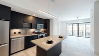 Located at 5211 Peachtree Blvd in Chamblee, Georgia, this single-floor unit showcases a clean, contemporary design with light-toned hardwood flooring that flows throughout the living spaces. The open-concept kitchen is a standout feature, equipped with dark matte cabinetry that extends to the ceiling, a large central island with a neutral quartz countertop, and a full suite of stainless steel appliances including a gas range, built-in microwave, dishwasher, and French door refrigerator. Sleek linear pendant lighting suspended above the island adds a refined, modern touch. The kitchen transitions seamlessly into a bright and open living area, which is bathed in natural light thanks to expansive floor-to-ceiling windows and sliding glass doors that lead to a private balcony with exterior views. The bedroom offers a calm, neutral palette with a warm accent wall, light hardwood floors, and a built-in closet with sliding doors providing ample storage. The bathroom is finished in a minimalist style, featuring large-format white floor tiles, a dark vanity with a quartz countertop, and a frameless glass-enclosed walk-in shower. Together, these spaces deliver a cohesive, upscale aesthetic suited for modern urban living.
