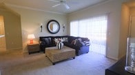 This single-floor residence offers a thoughtfully designed layout centered around a comfortable and inviting living area. The living room is finished with plush wall-to-wall carpeting underfoot and features elegant crown molding that frames the ceiling, lending a refined touch to the space. A ceiling fan is centrally mounted, providing both function and a subtle design element overhead. Large sliding glass doors dressed with vertical blinds span one wall, flooding the room with abundant natural light while offering access to the outdoors and views beyond. The warm, neutral-toned walls create a versatile backdrop that complements a range of interior styles. The room comfortably accommodates a large sectional sofa arrangement centered around a woven ottoman, with side tables providing convenient accent lighting. A decorative round mirror flanked by wall sconces serves as a focal point above the seating area, adding character and depth to the room. Overall, the space strikes a balance between casual comfort and tasteful design, making it well-suited for both everyday living and entertaining. The abundance of natural light and open feel contribute to a bright and welcoming ambiance throughout.