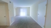 This single-floor space features an open living area finished with wall-to-wall carpeting in a neutral beige tone, providing a comfortable and cohesive foundation throughout the room. The walls are painted in a soft, light cream color, contributing to a clean and airy atmosphere. Natural light enters generously through a large sliding glass door fitted with vertical blinds, brightening the interior and creating a welcoming ambiance. The sliding door likely leads to an outdoor area, adding a sense of connectivity to the exterior. A standard entry door is positioned along one wall, offering convenient access to the unit. Overhead, the ceiling is smooth and uniform, maintaining the space's uncluttered aesthetic. Ventilation is provided via wall-mounted registers, ensuring practical climate control. The open layout of the living area allows for flexible furniture arrangement, making it adaptable to a variety of uses. Overall, the space presents a straightforward, functional design characterized by neutral finishes, ample natural light, and a simple, understated aesthetic well-suited for everyday living.