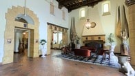 This remarkable single-floor space is a showcase of Spanish Colonial Revival architecture, exuding grandeur and historic character at every turn. The main hall commands attention with its soaring double-height ceilings, finished with dark, exposed wood beams and decorative painted accents that reflect a rich artistic tradition. Terracotta brick flooring runs throughout, lending warmth and an unmistakable old-world charm to the space.

Arched stone doorways frame the transitions between areas, their intricate carved detailing highlighting the exceptional craftsmanship of the structure. A large, ornately tiled fireplace serves as a commanding focal point along one wall, flanked by a richly upholstered seating arrangement featuring deep red velvet chairs and a tufted sofa set atop a classic patterned area rug. Tall sculptural elements and potted plants add organic dimension to the interior.

Natural light filters in through high clerestory windows, illuminating the white plaster walls and enhancing the sense of vertical space. A decorative hanging lantern provides ambient warmth overhead. Colorful tile friezes and ornamental details are woven throughout, reinforcing the space's cohesive and historically inspired aesthetic. Overall, the space offers a majestic, museum-quality ambiance that is both impressive and inviting.