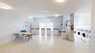 Located at 4415 Bathurst Street in Toronto, this shared laundry facility offers a well-organized and functional space designed to serve building residents efficiently. The single-floor layout is thoughtfully arranged, featuring distinct zones for washing, drying, and folding.

The main laundry area houses a row of front-loading washing machines and several stacked dryer units, providing ample capacity for multiple users. A card-operated payment kiosk is conveniently stationed near the entrance, supporting a cashless payment system. Natural light filters in through windows along one wall, keeping the space bright and pleasant, while a wall-mounted mini-split air conditioning unit ensures year-round comfort.

A dedicated utility area includes a stainless steel utility sink with a tile backsplash, ideal for pre-treating garments or hand-washing items. A separate folding station provides a large table with seating, offering residents a comfortable area to sort and fold laundry. Informational signage is posted throughout the space for easy reference.

The facility features durable terrazzo flooring throughout, easy to clean and well-suited to the demands of a high-traffic communal space. Light grey walls and white ceilings contribute to a clean, neutral aesthetic. Security is addressed with a windowed metal entry door and a wall-mounted camera. Overall, the space is practical, well-equipped, and maintained to a functional standard.