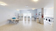 Located at 4415 Bathurst Street in Toronto, this shared laundry facility offers a well-organized and functional space designed to serve building residents efficiently. The single-floor layout is thoughtfully arranged, featuring distinct zones for washing, drying, and folding.

The main laundry area houses a row of front-loading washing machines and several stacked dryer units, providing ample capacity for multiple users. A card-operated payment kiosk is conveniently stationed near the entrance, supporting a cashless payment system. Natural light filters in through windows along one wall, keeping the space bright and pleasant, while a wall-mounted mini-split air conditioning unit ensures year-round comfort.

A dedicated utility area includes a stainless steel utility sink with a tile backsplash, ideal for pre-treating garments or hand-washing items. A separate folding station provides a large table with seating, offering residents a comfortable area to sort and fold laundry. Informational signage is posted throughout the space for easy reference.

The facility features durable terrazzo flooring throughout, easy to clean and well-suited to the demands of a high-traffic communal space. Light grey walls and white ceilings contribute to a clean, neutral aesthetic. Security is addressed with a windowed metal entry door and a wall-mounted camera. Overall, the space is practical, well-equipped, and maintained to a functional standard.