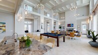 Located on Cimarron Boulevard in Corpus Christi, Texas, this single-floor community clubhouse is a thoughtfully designed gathering space that combines elegance with functionality. The interior is immediately striking, defined by soaring double-height ceilings accented with coffered detailing and an abundance of natural light streaming through oversized windows and transom glass panels.

The main gathering area serves as the heart of the space, featuring a full-size billiards table as a central focal point, surrounded by stylish rattan dining chairs and marble-topped tables. Warm chandelier lighting and sleek pendant fixtures illuminate the open-plan layout, which flows seamlessly across light-toned tile flooring throughout.

A dedicated lounge area offers a comfortable retreat, anchored by a dramatic floor-to-ceiling stacked stone fireplace with a bold stone hearth surround and a wall-mounted television above. Plush seating, navy blue accent rugs, and large windows with sheer drapery overlooking the outdoor pool area create a relaxed yet refined atmosphere.

The clubhouse also includes a well-appointed kitchen outfitted with crisp white shaker cabinetry, granite countertops, a white subway tile backsplash, stainless steel appliances, and a professional-grade range hood. A workstation area with exterior-facing windows provides additional functional space. The overall ambiance is coastal-contemporary, blending airy architecture with upscale finishes throughout.