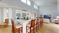 Located in Orange Beach, Alabama, this single-floor community space offers a welcoming, coastal-inspired atmosphere designed for comfort and gathering. The layout is open and expansive, with soaring vaulted ceilings and a bank of clerestory windows that flood the interior with natural light throughout the day. Large tile flooring in warm sandy tones runs continuously through the space, reinforcing the beachy aesthetic.

A central focal point is the elongated marble-topped bar counter, flanked by elegant wood-and-white upholstered bar stools — ideal for socializing or casual seating. The kitchen and bar area is well-appointed with recessed lighting and overlooks the main living area seamlessly.

The living room features a relaxed coastal décor with neutral upholstered sofas, teal accent chairs, woven rattan furnishings, and nautical-themed artwork, including a charming fish wall piece. A built-in bookshelf adds a cozy reading nook element to one corner, complemented by comfortable seating arrangements.

A dedicated workspace with a white desk and classic office chair provides a functional area near the entrance. White wainscoting and soft gray walls lend a clean, polished feel throughout. Ceiling fans ensure comfort year-round. Multiple glass-paned doors open to what appears to be an outdoor pool area, enhancing the indoor-outdoor connection.