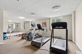 Located at 350 Elliott St in Windsor, Ontario, this fitness facility offers a practical and well-organized space dedicated to health and wellness. The exercise room serves as the centerpiece, featuring a wide variety of cardio equipment including treadmills, elliptical trainers, and a stationary bike, alongside strength-training machines and a mini trampoline. A wall-mounted television provides entertainment during workouts, while informational fitness posters — covering topics such as resistance training and heart rate guidelines — are displayed throughout for reference. Large mirrors along one wall enhance the sense of space and allow users to monitor their form. The room is finished with neutral carpet flooring and textured ceilings, with flush-mount lighting providing consistent illumination. Natural light enters through windows, contributing to a bright and functional atmosphere. A connecting hallway with tile flooring leads to additional amenities, most notably a wood-paneled sauna room that adds a relaxation element to the facility. The hallway also connects to storage areas via white panel doors. The layout flows logically from the main exercise area through the transitional hallway to the sauna, making the space efficient and easy to navigate for users seeking a comprehensive fitness experience.