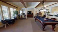 This well-appointed single-floor recreation and entertainment space is designed to accommodate a variety of social activities in a refined, comfortable setting. The main open area is anchored by a full-size billiard table positioned beneath an ornate wrought-iron pendant light fixture, setting an elegant tone throughout the space. The coffered ceiling, detailed with dark wood beams and recessed lighting, adds architectural character and a sense of generous vertical space.

The room is organized into distinct functional zones that flow naturally into one another. A lounge area features plush, warm-toned seating arranged around a large flat-screen television mounted above a media console flanked by matching bookshelves stocked with books and media. Bold framed artwork adorns the warm yellow walls, contributing to the inviting atmosphere. A secondary seating nook provides additional comfortable chairs beside large windows with plantation shutters that admit natural light.

Tall bistro-style tables with swivel stools offer casual seating near the windows, while individual computer workstations line one wall. Toward the rear, a kitchenette with dark cabinetry and appliances supports entertaining needs, with a door providing direct outdoor access. Potted greenery and decorative accessories are distributed throughout, enhancing the welcoming, community-oriented ambiance of the space.