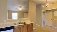 This single-story space showcases an open-concept layout that connects the kitchen to the adjoining living areas, creating a cohesive and functional flow throughout. The kitchen features warm oak cabinetry with raised panel doors, a double-basin stainless steel sink set into a light-colored countertop, and a pass-through opening that overlooks the adjacent living space. This architectural detail enhances the sense of openness and allows natural light from the living area window to filter through.

The neutral beige walls and light carpeting in the living and adjoining areas contribute to a consistent, understated palette throughout the space. A ceiling fan with an integrated light fixture is centrally positioned in the living area, providing both air circulation and ambient lighting. A wall-mounted light fixture adds additional illumination near the hallway leading to further rooms.

The hallway visible from the kitchen connects to additional rooms, hinting at a well-organized floor plan. A ceiling-mounted HVAC vent is visible, indicating a central air system. Overall, the space presents a straightforward and practical layout with traditional design elements suitable for comfortable everyday living.