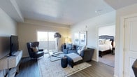 This single-floor residence showcases a thoughtfully designed living space that balances comfort and functionality. The living room serves as the central gathering area, finished with warm hardwood-style flooring that extends throughout the unit. A large sliding glass door floods the space with natural light and provides direct access to a private balcony, offering outdoor views that enhance the room's open feel. The textured ceiling adds subtle architectural character, while neutral wall tones create a versatile backdrop for various décor styles.

The living area is generously proportioned, comfortably accommodating a sectional sofa with a chaise, an accent armchair, a glass coffee table, and additional decorative elements, all anchored by a classic area rug. Framed wall art adds visual interest without overwhelming the clean, understated aesthetic.

An open doorway connects the living room directly to the bedroom, which features carpeted flooring, a substantial bed frame, and ample natural light from its own window. The seamless flow between the two rooms contributes to the unit's cohesive, well-organized layout, making the space feel both inviting and practical for everyday living.