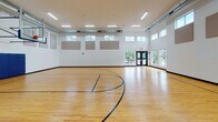 Located at 15655 Greythorne Dr in Charlotte, NC, this single-story recreational facility is purpose-built around a full indoor basketball court. The space is defined by its generous ceiling height, which accommodates regulation glass backboards and hoops at each end of the court. The polished hardwood floor is finished with clearly marked court lines in blue and black, providing a well-organized playing surface suitable for competitive and recreational use alike.

The interior walls are constructed of painted concrete block and feature strategically placed acoustic panels that help manage sound within the expansive space. Rows of clerestory windows along the upper walls admit natural light from multiple sides, complementing the flush-mounted linear fluorescent light fixtures overhead. Exposed ductwork runs along the ceiling, contributing to the facility's functional, utilitarian aesthetic.

Blue padded wall guards are installed along the lower portions of the end walls, providing player safety near the baskets. Double entry doors on opposing ends of the court offer convenient access and additional views to the surrounding exterior. The overall environment is clean, well-lit, and efficiently designed for athletic activity.