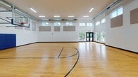 Located at 15655 Greythorne Dr in Charlotte, NC, this single-story recreational facility is purpose-built around a full indoor basketball court. The space is defined by its generous ceiling height, which accommodates regulation glass backboards and hoops at each end of the court. The polished hardwood floor is finished with clearly marked court lines in blue and black, providing a well-organized playing surface suitable for competitive and recreational use alike.

The interior walls are constructed of painted concrete block and feature strategically placed acoustic panels that help manage sound within the expansive space. Rows of clerestory windows along the upper walls admit natural light from multiple sides, complementing the flush-mounted linear fluorescent light fixtures overhead. Exposed ductwork runs along the ceiling, contributing to the facility's functional, utilitarian aesthetic.

Blue padded wall guards are installed along the lower portions of the end walls, providing player safety near the baskets. Double entry doors on opposing ends of the court offer convenient access and additional views to the surrounding exterior. The overall environment is clean, well-lit, and efficiently designed for athletic activity.