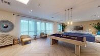 Located at 5475 N University in Lauderhill, Florida, this expansive single-floor amenity space offers a sophisticated and versatile environment designed for both leisure and socializing. The open-concept layout spans a generous footprint, unified by striking light-toned herringbone hardwood flooring that runs throughout, lending the space a refined yet approachable aesthetic.

A central highlight is the full-sized billiard table, positioned beneath a cluster of elegant gold pendant lights that add a warm, contemporary focal point. Adjacent to the game area, a comfortable lounge zone features a deep-blue sectional sofa, a travertine coffee table, and lush tropical plants set against a warm sand-toned accent wall with a large flat-screen television and a textured white media console.

Toward the entry, a welcoming reception-style area includes a navy-blue island bar with rattan bar stools, illuminated by a statement chandelier. This zone transitions seamlessly into casual seating clusters with light wood armchairs and side tables, perfect for informal gatherings.

Floor-to-ceiling windows draped in sheer white curtains line one entire wall, flooding the space with abundant natural light and offering views of the surrounding greenery. Decorative accents — including a sunburst wall mirror, botanical artwork, and indoor plants — complete the coastal-inspired, resort-style ambiance throughout.