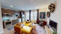 This single-floor unit offers a well-appointed open-concept layout that seamlessly connects the kitchen, living, and dining areas. The kitchen is outfitted with rich dark wood cabinetry, white quartz countertops, and a full suite of stainless steel appliances including a gas range, built-in microwave, French door refrigerator, and dishwasher. Light wood-style flooring runs throughout the main living area, complementing the bright, neutral walls and recessed lighting overhead. Large windows dressed with elegant draping curtains invite abundant natural light into the living space, where a bold mustard yellow sectional sofa anchors the room over a vibrant patterned area rug. A compact dining nook with a round black table and deep red chairs adds a stylish contrast. Decorative woven wall baskets and curated artwork contribute to the eclectic, globally inspired aesthetic. The bedroom features a sliding barn door, a woven rattan headboard, matching nightstands, and a textile wall hanging that adds warmth and character. The bathroom is finished with eye-catching patterned floor tile, a soaking tub with subway tile surround, built-in shelving, and elegant neutral tones throughout.