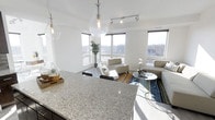 This contemporary single-floor space showcases a thoughtfully designed open-concept layout that seamlessly connects the kitchen and living areas. The kitchen is anchored by a generous granite countertop with striking speckled detailing, complemented by elegant glass pendant lights that hang at varying heights, adding a sculptural quality to the space. Track lighting runs along the ceiling, ensuring the entire area is well illuminated. The living area is bathed in natural light, courtesy of a series of large, floor-to-ceiling windows that wrap around the space and offer expansive views of the surrounding neighborhood and treeline. White walls and light-toned flooring contribute to a clean, airy aesthetic throughout. The living space is furnished with contemporary neutral-toned seating, a glass accent table, and a colorful area rug that adds warmth and visual interest. A tall potted plant near the windows brings a touch of greenery indoors. The overall ambiance is bright, modern, and inviting, with a cohesive design language that balances minimalist architecture with carefully chosen finishing touches to create a polished, comfortable environment.