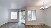 This single-floor space offers a clean, modern aesthetic defined by its abundance of natural light and open feel. The main living area features wood-look plank flooring that runs throughout, providing a warm yet contemporary foundation. Crisp white walls and smooth ceilings contribute to the bright, airy atmosphere, creating a neutral canvas well-suited to a variety of interior styles.

A prominent sliding glass door serves as a central architectural feature, framed by vertical blinds and offering direct views of mature surrounding greenery. Flanking the sliding door is a large picture window, also fitted with horizontal blinds, which further amplifies the natural light flowing into the space. An additional window on the adjacent wall ensures the room remains well-lit throughout the day.

Clean white baseboards trim the walls, adding a polished, finished look to the interior. The overall ambiance is calm and understated, with a move-in-ready condition and a layout that emphasizes openness and connection to the outdoors. The greenery visible through the windows adds a pleasant natural backdrop to the living environment.