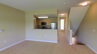 This two-story home offers a thoughtfully designed layout across its main and upper levels. The main level centers around an open-concept living area finished with neutral-toned carpeting and warm beige walls, creating a clean and inviting atmosphere. A pass-through kitchen counter visually connects the kitchen to the living space, making the floor plan feel open and functional. The kitchen is equipped with stainless steel appliances, including a refrigerator and microwave, complemented by light wood cabinetry and granite-style countertops. Recessed and overhead lighting illuminates the kitchen area effectively. A hallway beyond the kitchen leads toward a windowed entry door, allowing natural light to filter through the main level. A staircase with a white-painted banister is visible to the right, providing access to the upper floor. The overall aesthetic throughout the main level is understated and practical, with consistent neutral tones, clean lines, and a layout designed to maximize usable space. The upper floor offers additional living or sleeping accommodations, completing this well-organized multi-level home.