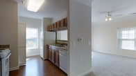 This single-floor residence offers a practical and well-organized layout, connecting key living areas through an open, flowing design. A carpeted hallway leads from the entry toward the main living spaces, framed by clean white walls and a six-panel door with brass hardware. Crown molding details add a subtle architectural touch throughout.

The kitchen is efficiently arranged and open to the adjacent living area, featuring rich medium-toned wood cabinetry that extends from base to upper wall level, providing ample storage. White appliances — including a refrigerator, dishwasher, and range — complement the neutral countertops. A double sink is positioned beneath upper cabinetry, and a window allows natural light to filter in. The kitchen floor is finished in hardwood, which contrasts with the carpeted areas of the adjoining spaces.

The living area flows naturally from the kitchen, finished with wall-to-wall carpet and a ceiling fan, contributing to a comfortable and airy atmosphere. The open connection between rooms enhances the overall sense of space. Neutral wall tones and consistent trim work throughout give the residence a cohesive, understated character.