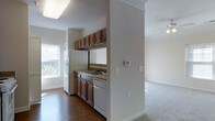 This single-floor residence offers a practical and well-organized layout, connecting key living areas through an open, flowing design. A carpeted hallway leads from the entry toward the main living spaces, framed by clean white walls and a six-panel door with brass hardware. Crown molding details add a subtle architectural touch throughout.

The kitchen is efficiently arranged and open to the adjacent living area, featuring rich medium-toned wood cabinetry that extends from base to upper wall level, providing ample storage. White appliances — including a refrigerator, dishwasher, and range — complement the neutral countertops. A double sink is positioned beneath upper cabinetry, and a window allows natural light to filter in. The kitchen floor is finished in hardwood, which contrasts with the carpeted areas of the adjoining spaces.

The living area flows naturally from the kitchen, finished with wall-to-wall carpet and a ceiling fan, contributing to a comfortable and airy atmosphere. The open connection between rooms enhances the overall sense of space. Neutral wall tones and consistent trim work throughout give the residence a cohesive, understated character.