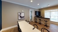 This single-floor space is thoughtfully designed to serve as a multifunctional communal area, blending dining, lounge, and kitchenette zones into a cohesive and welcoming environment. The interior is finished with consistent tile flooring and crisp white crown molding throughout, lending a polished, unified aesthetic.

The dining area features a long white table surrounded by classic black bentwood chairs, positioned beneath recessed lighting. A wall-mounted flat-screen television is installed above a bold teal accent wall, adding a striking focal point to the space. Abstract artwork is displayed on the neutral taupe walls, contributing to the contemporary character of the room.

The kitchenette is well-appointed with dark cabinetry, a black countertop peninsula, a stainless steel sink, microwave, and additional counter space suited for light food and beverage preparation. Bar-height seating along a secondary counter provides an additional casual dining option.

A separate lounge nook offers upholstered accent chairs in gray with nailhead trim, arranged around a small gold side table beneath a large framed artwork. Natural light enters through sizable windows fitted with blinds. A glass-paneled interior door connects the areas, enhancing the open, airy flow of the overall space.