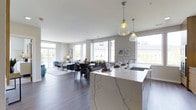 This elegant single-floor residence showcases a seamlessly integrated open-concept layout that connects the kitchen, dining, and living areas into one cohesive, flowing space. At the heart of the home is a striking kitchen island clad in white marble with bold grey veining, complemented by a sleek integrated sink and pendant lighting featuring delicate glass fixtures that cast a warm, inviting glow. The expansive open-plan living and dining area is bathed in natural light, courtesy of a generous array of large, floor-to-ceiling windows that frame views of the surrounding neighborhood. Rich hardwood flooring runs throughout, adding warmth and continuity to the contemporary interior. The living area is thoughtfully appointed with comfortable seating and tasteful decor, while the dining space accommodates both intimate gatherings and larger entertaining. Neutral wall tones and crisp white ceilings amplify the sense of openness, while carefully curated artwork and accessories lend a refined, polished character to the space. The overall ambiance is one of sophisticated modern living — airy, light-filled, and meticulously designed for both comfort and style.