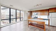 This contemporary single-floor residence showcases a thoughtfully designed open-concept layout that seamlessly connects the living, dining, and kitchen areas. Expansive floor-to-ceiling windows wrap around the main living space, flooding the interior with natural light and offering sweeping views of surrounding greenery and the urban streetscape beyond.

The kitchen is a standout feature, equipped with rich warm-toned wood cabinetry, stainless steel appliances including a range, microwave, and refrigerator, a white subway tile backsplash, and a granite-topped island that provides additional prep and storage space. A stylish sliding barn door adds a modern architectural detail at the kitchen's edge.

The generously sized bedrooms feature plush carpeting and large windows that continue the theme of natural light throughout the unit. One bedroom includes an en-suite bathroom and a walk-in closet. The bathroom is finished with classic white subway tile surrounding a full soaking tub, a wood-toned vanity, and light stone-look floor tiles.

Throughout the main living areas, light wood-grain flooring ties the space together, complementing the clean white walls and contemporary track lighting. The overall ambiance is bright, open, and modern, with a neutral palette that offers a versatile canvas for any interior style.