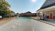 Located at 1980 W Main St in Central, South Carolina, this outdoor recreational area centers around a generously sized swimming pool with crystal-clear blue water. The pool deck offers ample space for lounging, lined with multiple sun chairs positioned to take advantage of the open sky and natural surroundings. Mature trees and flowering shrubs frame the perimeter, providing a lush, park-like backdrop that enhances the sense of privacy and tranquility.

A ping-pong table is conveniently situated on the deck near the pool, adding a recreational element beyond swimming. The entire pool area is enclosed by a classic wrought iron fence, ensuring both security and an open, airy aesthetic.

Adjacent to the pool is a single-story facility building constructed with a combination of lap siding and stacked stone veneer accents, giving it a clean, modern-rustic character. Vibrant orange doors mark the entrances to the restroom and changing facilities, while a covered porch area provides shade and a transition space between the indoors and the pool deck. A drinking fountain is accessible along the exterior wall.

The surrounding community is visible in the background, with residential buildings nestled among mature landscaping. The overall ambiance is bright, welcoming, and well-maintained, making this an ideal outdoor amenity space for warm-weather recreation.