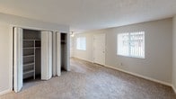 This single-story unit is thoughtfully laid out across a compact yet efficient floor plan. The kitchen is equipped with warm medium-toned wood cabinetry, light stone-look laminate countertops, and black appliances including a full-size refrigerator. A pass-through opening connects the kitchen to the adjacent living area, which features smooth wood-look flooring and a ceiling fan with integrated lighting. A window at the far end of the living space admits natural light and provides views to the exterior.

The living area transitions seamlessly into carpeted spaces, including a generously sized main room with a textured ceiling, two windows fitted with vertical blinds, and a built-in closet with bi-fold doors offering organized storage. Additional carpeted rooms provide flexible use as bedrooms or multipurpose spaces. Each room maintains a neutral color palette of soft grays and whites, contributing to a clean, understated aesthetic throughout.

The bathroom includes a full tub-shower combination with tile surround, a toilet, and a vanity with a stone-look countertop, finished with neutral gray walls. Throughout the unit, consistent use of light-toned walls, textured ceilings, and white trim creates a cohesive and practical living environment.
