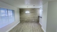 This single-story space showcases a thoughtfully designed interior with a blend of modern and rustic elements. The dining area serves as a focal point, highlighted by a full wood-plank accent wall that adds warmth and texture to the room. Luxury vinyl plank flooring in a neutral gray-toned wood finish extends throughout, creating a cohesive and contemporary look underfoot.

A elegant chandelier with globe lights hangs above the dining area, providing a decorative centerpiece and ambient illumination. An additional pendant light is visible nearby, suggesting a transition into an adjacent kitchen or bar area, where a dark countertop offers a striking contrast to the lighter tones of the space.

The interior is finished with clean white walls and white baseboards, contributing to a bright and airy atmosphere. A window fitted with horizontal blinds allows for natural light and privacy control. The textured popcorn ceiling adds a classic touch overhead. The open layout between the dining and kitchen areas promotes a functional flow, making the space well-suited for everyday living and entertaining alike.