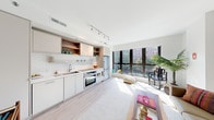 This single-floor residence showcases a thoughtfully designed open-plan layout that seamlessly connects the kitchen and living areas. The kitchen is outfitted with flat-front cabinetry in a warm neutral palette, open wood-accented shelving, classic white subway tile backsplash, and a full suite of stainless steel appliances, including a built-in oven, microwave, and refrigerator. Track lighting overhead ensures the workspace is well-illuminated.

The living area benefits from an abundance of natural light, courtesy of expansive floor-to-ceiling windows that frame views of surrounding greenery. Light hardwood flooring runs throughout the main living spaces, enhancing the bright, airy ambiance. A hallway connects the public areas to the private quarters, maintaining a clear and functional flow.

The bedroom is a calm retreat, featuring plush carpeting, a tray ceiling detail, and a sliding closet. The bathroom is finished with large-format slate-toned floor tiles, a floating wood-grain vanity with an integrated sink, a framed mirror cabinet, and a full tub-shower combination. Clean lines and a restrained material palette lend the entire residence a cohesive, contemporary character.