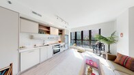 This single-floor residence showcases a thoughtfully designed open-plan layout that seamlessly connects the kitchen and living areas. The kitchen is outfitted with flat-front cabinetry in a warm neutral palette, open wood-accented shelving, classic white subway tile backsplash, and a full suite of stainless steel appliances, including a built-in oven, microwave, and refrigerator. Track lighting overhead ensures the workspace is well-illuminated.

The living area benefits from an abundance of natural light, courtesy of expansive floor-to-ceiling windows that frame views of surrounding greenery. Light hardwood flooring runs throughout the main living spaces, enhancing the bright, airy ambiance. A hallway connects the public areas to the private quarters, maintaining a clear and functional flow.

The bedroom is a calm retreat, featuring plush carpeting, a tray ceiling detail, and a sliding closet. The bathroom is finished with large-format slate-toned floor tiles, a floating wood-grain vanity with an integrated sink, a framed mirror cabinet, and a full tub-shower combination. Clean lines and a restrained material palette lend the entire residence a cohesive, contemporary character.