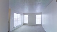 This single-floor space offers a clean, straightforward layout designed for comfort and functionality. The main room is generously proportioned, featuring soft wall-to-wall carpeting that adds warmth underfoot and a classic popcorn-textured ceiling that lends a familiar, residential feel overhead. Crisp white walls provide a neutral backdrop throughout, making the interior feel open and easy to personalize.

Natural light is a defining characteristic of this space. Three windows line the exterior wall, each dressed with horizontal blinds that allow for flexible light control. The windows look out toward a neighboring residential streetscape, offering a sense of connection to the surrounding community while still maintaining privacy.

The room's layout is open and unobstructed, providing a versatile footprint suitable for a variety of uses and furniture arrangements. White baseboards frame the perimeter of the room, adding a clean finishing detail that complements the overall neutral palette. The combination of abundant natural light, light-toned carpet, and bright walls creates an airy, welcoming ambiance throughout the interior.