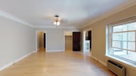 This well-appointed single-floor residence showcases a thoughtful blend of classic architectural details and functional design. The main living area is generously proportioned, featuring warm light hardwood floors that run throughout and elegant crown molding that frames the ceilings. A ceiling fan with a light fixture provides both illumination and airflow, while neutral gray-toned walls create a clean, versatile backdrop. Multiple doorways connect the living space seamlessly to the rest of the home, promoting an open and airy feel.

The kitchen is a standout feature, accessed through a distinctive arched entryway that adds a touch of architectural character. Inside, rich dark-wood cabinetry lines both walls in a galley-style layout, complemented by granite-look countertops and black appliances including a refrigerator, gas range, and microwave. Built-in display cabinets with glass panels flank the kitchen entrance, adding both charm and additional storage.

The bedroom is a comfortable, well-lit room with the same hardwood flooring and crown molding found throughout the home. A spacious walk-in closet with wire shelving and hanging rods offers ample storage. The bathroom features classic white subway tile surrounding a full-size soaking tub with an overhead shower, brightened by a natural light window.