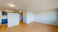 This single-floor unit offers a thoughtfully designed layout with light hardwood flooring that flows seamlessly throughout the space, creating a warm and cohesive aesthetic. The main living area is generously proportioned, bathed in abundant natural light streaming through a striking wall of wide panoramic windows that frame sweeping views of the surrounding cityscape and distant mountains — a standout feature of the space.

The galley-style kitchen is efficiently arranged and well-equipped, featuring slate-blue cabinetry, white countertops, stainless steel appliances including a full-size refrigerator and an electric range, and a single-basin sink. A window above the sink allows natural light to fill the kitchen while offering additional exterior views. The kitchen connects directly to the rest of the unit, providing an easy flow between cooking and living spaces.

Built-in shelving units provide practical storage within the living area, complementing the clean, minimalist design of the white walls and flush-mount ceiling lighting. The bathroom includes a full tub-shower combination finished with classic white subway tile, brushed gold hardware, and wood-tone flooring that ties into the overall design of the unit. The space presents a functional, well-maintained interior with a bright and open atmosphere throughout.