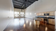 Located at 540 S Saginaw St in the heart of downtown Flint, MI, this two-story loft offers a striking blend of industrial character and modern finishes. The main floor commands attention with soaring ceilings that expose dramatic black steel trusses and ductwork overhead, while rich, dark-stained hardwood floors run throughout. Large windows flood the expansive open living area with natural light and frame views of the surrounding cityscape. The kitchen is a standout feature, centered around a sculptural curved island with sleek black granite countertops, light maple cabinetry, mosaic tile backsplash, pendant lighting, and stainless steel appliances. A modern staircase with a wood-and-metal cable railing leads to the upper level, which offers additional open living space beneath the exposed truss ceiling. The spacious primary bedroom features a generous built-in closet and clean white walls complemented by the continuous hardwood flooring. Two bathrooms are finished with tile flooring, warm maple vanities, and black granite countertops. A utility room and multiple hallways complete the thoughtful layout of this distinctive urban loft.