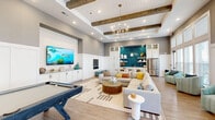 This thoughtfully designed amenity lounge delivers a spacious, open-plan layout that seamlessly blends entertainment, dining, and relaxation into one cohesive space. The room's elevated ceilings are accented by exposed rustic wood beams and a shiplap ceiling detail, lending warmth and character to the otherwise contemporary interior. Recessed lighting and a statement chandelier provide layered illumination throughout.

The entertainment zone anchors one end of the space, featuring a wall-mounted large-screen TV above custom white cabinetry, flanked by cue stick storage and a full-size pool table — ideal for social gatherings. Adjacent to this, a generous sectional sofa arranged around a round drum coffee table creates an inviting lounge area centered on a patterned area rug.

Toward the rear, a dining table with seating for several is positioned next to a well-appointed kitchenette. The kitchenette showcases a bold teal herringbone tile backsplash, open display shelving, white cabinetry, and a stainless steel refrigerator — a vibrant focal point in the space.

A sun-filled reading nook lines the windowed wall, where rows of floor-to-ceiling windows flood the area with natural light and outdoor views. Striped upholstered accent chairs are arranged here, offering a quiet retreat. Light hardwood flooring and white wainscoting unify the entire space with a clean, coastal-inspired aesthetic.