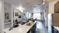 This thoughtfully designed single-floor residence showcases a seamless open-concept layout that connects the kitchen, dining, and living areas into one cohesive, inviting space. The kitchen serves as the heart of the home, featuring a generous island with a sleek white quartz countertop, a deep stainless steel sink, and dark cabinetry with gold-toned hardware. Elegant pendant lighting hangs above the island, adding a warm and contemporary touch. The dining area flows naturally from the kitchen, offering a comfortable setting for entertaining. The living room is bathed in natural light from a large picture window dressed with soft draping curtains, illuminating the wood-style flooring and neutral-toned walls accented with soft greens. Track lighting overhead provides additional ambiance throughout the space. A glimpse into the bedroom reveals a continuation of the modern aesthetic, with plush furnishings and coordinated décor. Throughout, the design balances functionality with style, creating a bright, airy atmosphere that feels both welcoming and refined. The consistent use of cool tones, quality finishes, and carefully curated details gives this residence a polished, move-in-ready appeal.