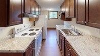 This single-floor unit offers a practical and comfortable layout with distinct living, sleeping, kitchen, and bathroom areas. The open living room is finished with warm hardwood-style flooring, neutral walls, and a textured ceiling, creating a bright and welcoming atmosphere. Natural light filters in through wood-trimmed windows, and a wall-mounted air conditioning unit and baseboard heating ensure year-round comfort. The galley-style kitchen is well-equipped, featuring rich dark wood cabinetry, light stone-finish countertops, a stainless steel sink, and white appliances including an electric range and refrigerator. A window at the end of the kitchen brings in additional natural light. The bedroom is carpeted for warmth and comfort, and includes a ceiling fan with light fixture, a window with an exterior view, baseboard heating, and a closet with louvered bifold doors. The bathroom is clean and functional, appointed with a full tub and shower combination surrounded by classic cream-toned tile walls and chrome fixtures. A small window with a curtain adds a touch of natural light. The entry hallway features a wood-paneled door, providing a defined and private entrance to the unit. Overall, the space presents a cohesive, move-in-ready environment with a neutral palette throughout.