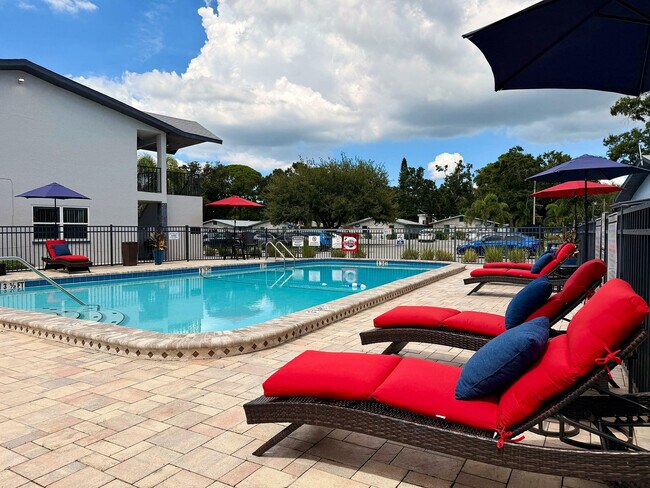 Acogedora zona de piscina en un complejo de apartamentos, con tumbonas y sombrillas de color rojo brillante bajo un cielo azul despejado. - Waterside Village