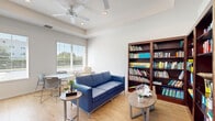 Located at 2115 Lepic Ln in Kissimmee, Florida, this single-room office space offers a thoughtfully designed, multi-functional environment suited for both focused work and relaxed collaboration. The space features light hardwood flooring throughout, crisp white walls, and an elevated tray ceiling with a ceiling fan that enhances the sense of openness and airflow.

A dedicated workstation area occupies one corner, outfitted with rich dark wood cabinetry overhead, dual monitors, and ergonomic seating for multiple users — providing an organized and productive work zone. Opposite the workstation, a generous bank of large windows floods the space with natural light while offering views of the surrounding exterior, creating a bright and welcoming atmosphere.

The lounge area is anchored by a bold blue sofa paired with round side tables, offering a comfortable setting for informal meetings or relaxation. An extensive built-in library wall with floor-to-ceiling dark wood bookshelves lines one section of the room, stocked with a wide variety of books and adding both character and intellectual warmth to the space.

Recessed lighting and ceiling-mounted fixtures complement the natural light, ensuring the space is well-lit throughout. The overall ambiance is clean, modern, and inviting — balancing professional functionality with a comfortable, community-oriented feel.