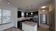 This single-floor space centers around a well-appointed kitchen that serves as the focal point of the layout. The kitchen features dark espresso-toned cabinetry that contrasts sharply against light-colored walls, creating a bold, contemporary aesthetic. Granite countertops with subtle veining extend across the workspace and onto a central island, which provides additional prep space and casual seating potential. Stainless steel appliances, including a refrigerator, electric range, built-in microwave, and dishwasher, complement the modern design scheme.

Overhead, a combination of track lighting and two sleek pendant lights hanging above the island ensures the kitchen is well-illuminated. A window above the sink introduces natural light into the space. The kitchen opens toward an adjoining area finished with neutral-toned carpet, suggesting a connected living or dining space. A corridor visible in the background hints at additional rooms beyond. The overall ambiance is clean, modern, and functional, with a cohesive dark-and-light contrast that gives the space a polished, intentional character.