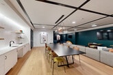 The Urban Unwind Room is a thoughtfully designed, single-floor amenity space that blends social, dining, and recreational functions into one cohesive environment. The main open area features light hardwood flooring, a distinctive coffered ceiling with bold black grid beams, and recessed lighting complemented by a sculptural pendant fixture. A large, dark-topped communal dining table surrounded by a mix of colorful mid-century-style chairs serves as a central gathering point, while a generously sized sectional sofa anchors an adjacent lounge zone. A wall-mounted television, deep teal accent walls, and curated artwork — including large-format paintings — contribute to the space's vibrant yet refined character. A built-in kitchenette with open shelving, white cabinetry, and a sink provides functional support for events and gatherings. A separate, dedicated room houses a TruGolf simulator, featuring synthetic turf hitting mats, a projection screen, and matte black walls that create an immersive, focused environment. Throughout the space, the design balances an urban-modern aesthetic with comfort-forward furnishings, making it well-suited for both casual relaxation and organized social activities.