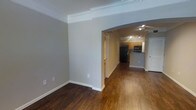 This single-floor residence offers a clean, well-appointed layout designed for comfortable everyday living. The main living area is finished with rich, dark hardwood flooring that extends throughout the space, creating a warm and cohesive aesthetic. Crown molding lines the ceiling, adding a refined architectural detail that elevates the overall feel of the interior. Neutral gray walls provide a versatile backdrop throughout the home.

An arched passageway connects the living area to the rest of the unit, lending a subtle architectural character to the open layout. Beyond the archway, a hallway leads to a fully equipped kitchen, which features warm wood-toned cabinetry, a raised breakfast bar or counter overhang ideal for casual dining, and a suite of black appliances including a refrigerator and microwave. Track lighting illuminates the kitchen, ensuring ample light for meal preparation.

A closet with louvered doors is visible near the kitchen area, offering practical storage solutions. The transition between the living and kitchen spaces flows naturally, making the layout feel connected and easy to navigate. Overall, the space presents a tidy, move-in-ready interior that balances functional design with tasteful finishing touches.