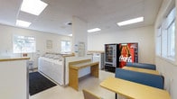 This single-floor laundry facility is designed with practicality and comfort in mind, offering a well-organized space for residents or patrons to complete their laundry needs efficiently. The room is brightly illuminated by recessed panel lighting set into a drop ceiling, creating a clean and welcoming atmosphere throughout.

A row of top-load washing machines lines one side of the room, accompanied by dedicated folding counters with a warm wood-toned finish and white cabinetry below, providing ample surface area for sorting and folding laundry. Informational notices and instructions are posted on the walls, keeping users well-informed about machine operation and facility guidelines.

Natural light filters in through windows along the exterior wall, adding to the overall brightness of the space. Vending machines, including a snack dispenser and a prominent Coca-Cola beverage machine, are positioned along one wall, offering convenient refreshment options while waiting. A casual seating and table area with light wood tables and blue chairs is situated toward the front of the room, giving patrons a comfortable place to relax during their visit. The flooring throughout is a durable, easy-to-clean surface suited to the demands of a high-traffic utility space. Overall, the facility is clean, functional, and thoughtfully arranged.